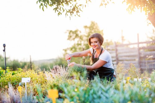 A woman with dark hair tied back, wearing a white t-shirt and dark gardening apron, is kneeling in a lush garden with a variety of plants and flowers. She is smiling as she tends to the garden bed, which includes green foliage and blooming flowers in shades of purple, yellow, and white. The garden is bordered by a wooden fence and features a well-maintained lawn area with dense, healthy grass. In the background, there are trees, shrubs, and a soft sunlight filtering through the branches, suggesting a warm, sunny day. The scene reflects a well-organised outdoor space suitable for gardening and landscaping services, with attention to plant care and garden aesthetics typical of properties in Whitechapel, London, and the surrounding areas. The overall environment is vibrant, healthy, and inviting, emphasizing professional gardening and outdoor maintenance aimed at enhancing curb appeal and outdoor enjoyment.