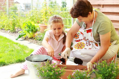 A woman and a young girl are engaged in gardening activities in a well-maintained outdoor space, with the woman wearing a colorful floral apron and gloves, and the girl dressed in a light pink top and pink striped trousers. They are working together in a raised flower bed, which contains dark, rich soil, and are handling gardening tools, possibly planting or tending to plants. The background showcases a lush garden with vibrant green foliage, a wire fence, and various potted plants, indicating a private residential garden in Whitechapel. The garden features neatly arranged flower beds, a paved area for gardening, and garden pots with flowering plants, creating a peaceful and inviting environment. The weather appears sunny and pleasant, enhancing the greenery and natural tones of the garden, which is managed by local gardening services such as Landscapers Whitechapel to maintain its tidy and vibrant appearance.