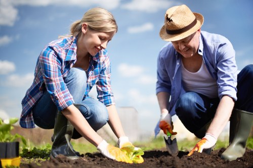 A young woman and an older woman are working together outdoors in a garden, planting or tending to the soil. The younger woman has blonde hair tied back and is wearing a blue and red plaid shirt with blue jeans, while the older woman is wearing a straw hat, a light blue jacket, and dark trousers. Both are wearing gardening gloves and are focused on their task, using small garden tools to handle the soil. The garden features dark, freshly turned earth, with a few green plants visible in the foreground. They are positioned on a well-maintained outdoor lawn, under a partly cloudy sky, suggesting a mild, sunny day. The background hints at other garden areas with additional planting beds. This scene illustrates typical gardening activities suitable for professional landscaping or home garden maintenance, reflecting the services offered by Landscapers Whitechapel in local outdoor environments near Whitechapel, London, aligning with gardening and landscaping service SEO signals.