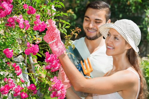 A man and a woman are gardening together outdoors in a lush garden, with vibrant pink flowering bushes and green foliage surrounding them. The woman, wearing a white sun hat and gardening gloves, is reaching up to tend to the flowering shrub, while the man, dressed in a light-colored t-shirt, is holding a small garden fork and smiling. The garden features a mix of healthy green grass, neatly trimmed hedges, and flowering plants, indicating an actively maintained outdoor space suitable for gardening services. Bright natural daylight highlights the vivid colours of the flowers and the greenery, creating a welcoming and well-kept garden environment. The scene captures a moment of outdoor maintenance and gardening care, typical for landscaped outdoor areas in Whitechapel or nearby London suburbs, and reflects the professional gardening services provided by Landscapers Whitechapel.