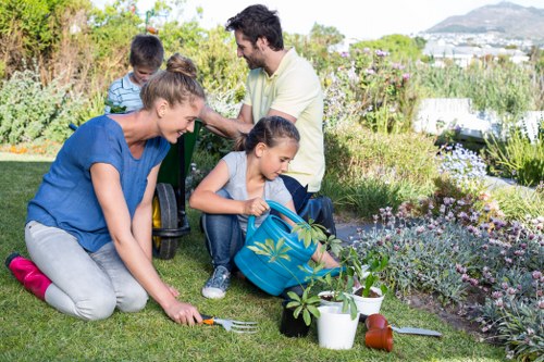 A family of four enjoying outdoor gardening in a well-maintained backyard, with a lush green lawn, flower beds with blooming plants, and various garden tools and potted plants arranged in the foreground. The scene features a woman kneeling on the grass, planting or tending to a small shrub, while a young girl helps by holding a watering can nearby. In the background, a man and a young boy, both seated on the grass, observe and participate in the gardening activity. The garden is bordered by dense shrubbery and ornamental plants, with a scenic view of distant hills and a partly cloudy sky, suggesting a pleasant weather day in Whitechapel. This image showcases family involvement in garden maintenance, emphasizing the natural textures of grass, soil, and flowering plants, and reflecting outdoor gardening and landscaping activities typical of the local area, which could be supported by services from Landscapers Whitechapel.