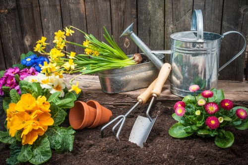 In the outdoor garden area of a residential property in Whitechapel, a section of freshly turned dark brown soil is prepared for planting, with gardening tools such as a garden fork, trowel, and watering can placed nearby. To the left, there are vibrant flowering plants, including yellow primroses with bright blooms and green foliage, as well as purple and pink flowering plants with rounded flowers, adding natural colour to the scene. An upside-down terracotta plant pot lies on the soil, indicating ongoing gardening work. The garden's background features a weathered wooden fence, providing privacy and a rustic backdrop to the landscaped outdoor space. The scene is well-lit, suggesting it is a clear, sunny day, ideal for gardening activities. This setting illustrates typical lawn and garden maintenance practices, involving planting, soil preparation, and caring for flowering plants, aligned with professional gardening services offered by Landscapers Whitechapel in the local Whitechapel area, postcode E1, supporting a well-kept and vibrant garden environment.