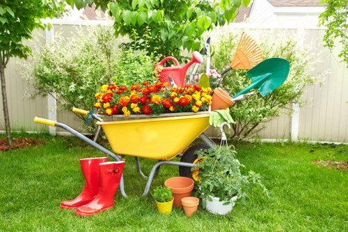 This image depicts a well-maintained residential garden space with a lush, green lawn bordered by a white wooden fence. At the centre, there is a colourful wheelbarrow filled with vibrant red and yellow flowering plants, surrounded by gardening tools and accessories including a red watering can, a pair of red rubber boots, several small flower pots, and a white planter with green foliage. In the background, leafy trees and dense shrubbery provide natural shade and privacy, creating a peaceful outdoor environment. The scene is set in bright daylight with clear weather, highlighting the natural tones of the grass, plants, and garden features. Such garden elements and maintenance tools suggest the presence of professional gardening and landscaping services, like those offered by Landscapers Whitechapel, who specialise in outdoor landscape enhancements and garden care in the Whitechapel area, inferred from the postcode and local context. This setting exemplifies a typical UK garden ready for seasonal planting and ongoing outdoor upkeep, contributing to the overall appeal of a landscaped outdoor space.
