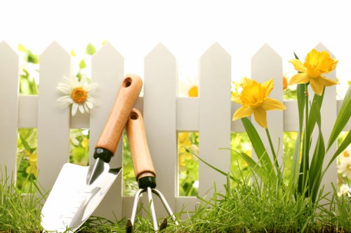 The image shows a small section of a well-maintained garden with a white picket fence in the background, featuring a few bright yellow daffodils and white daisies growing beside it. In the foreground, there is a lush, green lawn with dense grass that appears healthy and neatly trimmed. Two gardening tools— a hand trowel and a small fork with light wooden handles—are resting on the grass, suggesting ongoing gardening or lawn care activity. The scene is outdoors, illuminated by natural daylight, indicating fair weather conditions. This setting reflects typical features of a residential garden space in Whitechapel or similar areas, emphasizing the importance of lawn and flower bed maintenance which landscaping services like those offered by Landscapers Whitechapel often support to enhance outdoor curb appeal and garden health.