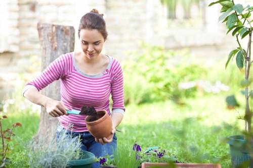 A young woman in a pink and white striped long-sleeve shirt is gardening outdoors in a residential garden, holding a small terracotta flower pot filled with soil and using a trowel to add soil to the pot. The garden features a well-maintained lawn with lush, green grass, bordered by various potted plants and flower beds containing purple and green foliage. In the background, there is a weathered wooden garden bench, a tree trunk, and a stone wall with some foliage, suggesting a private, landscaped outdoor space. The scene is illuminated by natural daylight, indicating a clear weather day, and the environment appears tidy and well-kept, highlighting professional gardening and lawn care services. The setting demonstrates an area suitable for garden planting and outdoor maintenance, consistent with local gardening services in Whitechapel or nearby areas.