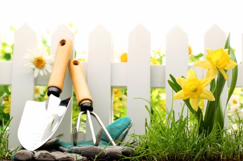 A garden scene in a landscaped outdoor space featuring a white picket fence in the background, with sunlight filtering through. In the foreground, there is a patch of lush, green grass with a few yellow daffodils in bloom growing near the fence. To the left, there are gardening tools including a small hand trowel with a wooden handle, a hand rake, and a pair of gardening gloves, all resting on the soil or on a surface. The garden area appears well-maintained with healthy plants, representative of typical gardening and lawn care activities performed by landscapers in Whitechapel or surrounding areas, providing a neat, vibrant, and inviting outdoor environment set during the daytime with clear weather.