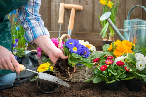 In the image, a person wearing a blue plaid shirt is planting a vibrant purple primula flower into dark, freshly turned soil within a garden bed. The surrounding area features various potted flowering plants with bright yellow, pink, and white blooms, arranged on the garden surface. There are gardening tools visible, including a small hand trowel in the person's right hand and a green watering can placed nearby, indicating active gardening work. The garden setting includes a wooden fence in the background, with a wooden garden panel and a watering can hanging, suggesting a well-maintained outdoor space. The soil appears rich and well-prepared, and the garden is reflected under natural light, likely on a clear day, with the overall scene emphasizing planting and outdoor horticultural activity suitable for homeowners in Whitechapel seeking professional gardening services from Landscapers Whitechapel.