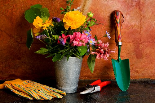 A close-up of a garden workspace showing a metal pot filled with a colourful arrangement of fresh flowers, including yellow, pink, and purple blooms, placed on a dark, textured surface against a warm-toned background. To the right of the pot, there is a small green-handled trowel with a red grip, and a pair of pruning shears with black and red handles. On the left side of the surface, a pair of yellow and green striped gardening gloves are neatly folded. The scene suggests outdoor gardening activity, with tools and flowers ready for planting or arranging, reflecting the gardening services offered by Landscapers Whitechapel, located in the nearby area, highlighting the importance of well-maintained outdoor spaces in the local community.