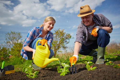 A woman and a man are working together in an outdoor garden in Whitechapel, engaging in planting young green seedlings into freshly tilled dark brown soil. The woman, with blonde hair and a blue checkered shirt, is smiling as she waters the small plants with a yellow watering can. The man, wearing a straw hat, plaid shirt, and green rubber boots, is crouched beside her, carefully tending to the plants with gardening gloves on, in a scene set under a partly cloudy sky. The garden features a clear, well-organized layout with a front section of lush grass, a boundary of low hedges, and several budding plants arranged in neat rows. In the background, there are trees and an open grassy area, emphasizing a peaceful outdoor environment suitable for gardening and landscaping activities. The natural tones of green, brown, and blue highlight the healthy growth of the garden space, supported by professional gardening services from Landscapers Whitechapel, with the overall scene suggesting a community or residential outdoor area in the postcode district surrounding E1.
