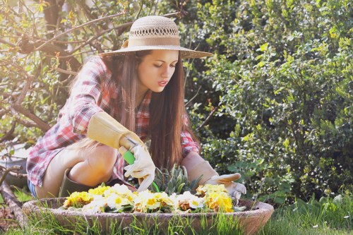 A young woman wearing a wide-brimmed straw hat, a plaid shirt, and gardening gloves is kneeling in a well-maintained garden during daytime, tending to a circular flower bed filled with various blooming plants that display yellow, white, and purple flowers. Behind her, dense greenery comprises a hedge or bush, creating a natural backdrop with shades of green from dark leaves to lighter foliage. The garden features a lush, evenly cut grass area surrounding the flower bed, with a few visible patches of soil or mulch at the edge of the planting area. Soft sunlight filters through the trees, casting gentle light on the scene, indicative of a clear, calm day suitable for outdoor gardening. The environment reflects a neat, landscaped outdoor space that could be part of a residential garden in Whitechapel or nearby in East London, aligning with professional gardening and landscaping standards provided by Landscapers Whitechapel, emphasizing natural beauty and outdoor upkeep.