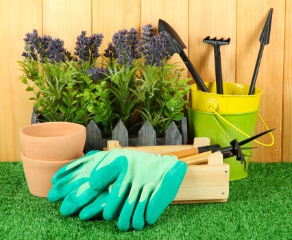 An outdoor gardening workspace on a landscaped area featuring a row of potted lavender plants with purple flower spikes and green foliage placed at the back. In front of the plants, there are several empty terracotta pots arranged on a surface covered with artificial green grass. To the right, a yellow plastic bucket holds various gardening tools such as a trowel, a hand rake, and a transplanter. A pair of teal gardening gloves lie on a light-colored wooden tool tray, which is positioned on the grass. The background consists of vertical wooden fencing, suggesting a private garden or backyard setting, with natural daylight illuminating the scene, indicating mild weather conditions suitable for outdoor gardening activities. The overall arrangement emphasizes tools and plant care items typical for garden maintenance and landscaping in Whitechapel or similar London areas, linking the image to professional gardening services offered by Landscapers Whitechapel.