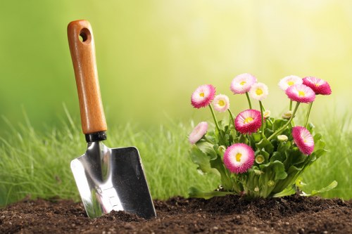 A close-up view of a small garden bed featuring a garden trowel with a wooden handle partially embedded in dark, freshly turned soil. To the right of the trowel, a cluster of pink and white daisy-like flowers with round, vibrant blooms and green foliage is visible, adding colour to the scene. The background shows a lush, green grassy area with a blurred effect, suggesting a well-maintained outdoor space in a residential garden, likely in Whitechapel or nearby in London. The overall setting appears well-organised and cared for, reflecting typical features of local gardening services provided in the area by companies like Landscapers Whitechapel. The natural daylight creates a bright and inviting atmosphere, highlighting the healthy plant growth and the readiness for planting or further garden maintenance.