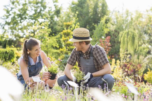 The image depicts a gardener and a young woman working together in a lush, landscaped garden located in Whitechapel. They are kneeling on a well-maintained lawn with dense, green grass, surrounded by vibrant flower beds filled with various blooming plants and small shrubs. In the background, there are mature trees with leafy branches providing dappled sunlight, creating a bright and inviting outdoor environment. The gardener, dressed in a checkered shirt, grey apron, and garden gloves, is holding a small potted plant, while the young woman, wearing casual attire and a denim apron, is smiling and engaging in planting or tending to the plants. The scene emphasizes a peaceful, well-organized outdoor space that showcases effective gardening techniques, with a focus on healthy plant growth, soil, and garden maintenance. This setting aligns with professional gardening and landscaping services offered by Landscapers Whitechapel, designed to enhance outdoor environments for residential properties in the area, including the postcode sector corresponding to Whitechapel.